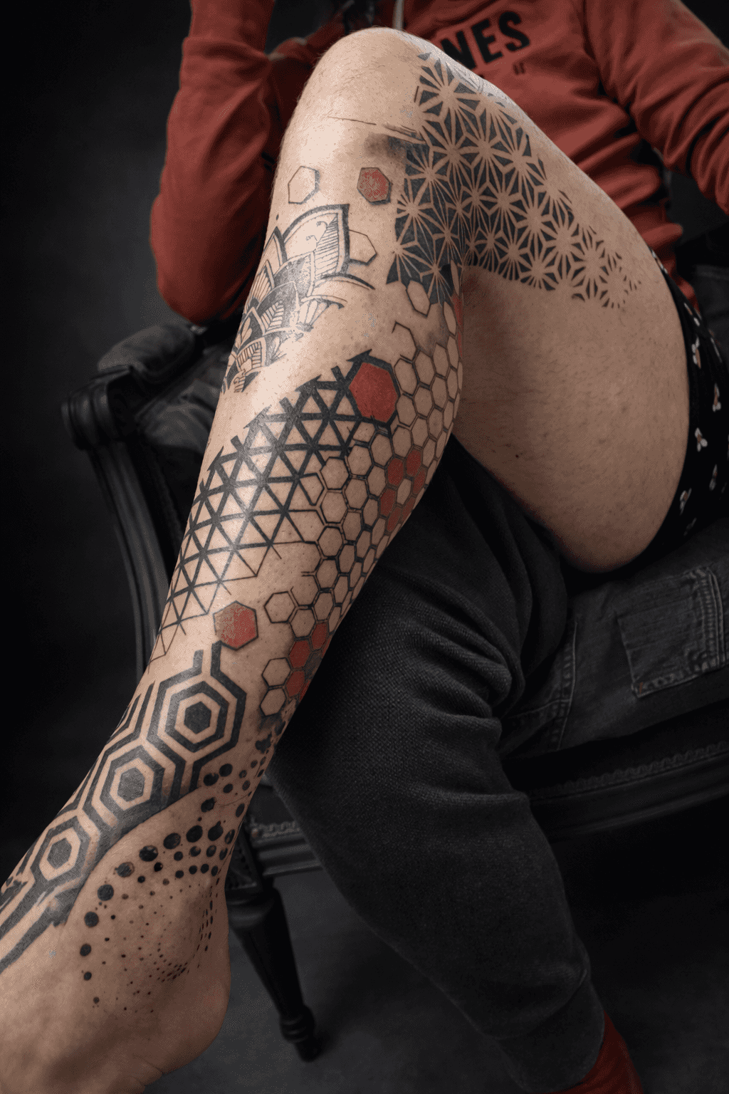 Personne assise avec la jambe ornée de motifs géométriques, composés d'hexagones et de points en noir et rouge, réalisés par Steven Chaudesaigues au Bastia Tattoo, salon de tatouage à Bastia, portant une chemise rouge.