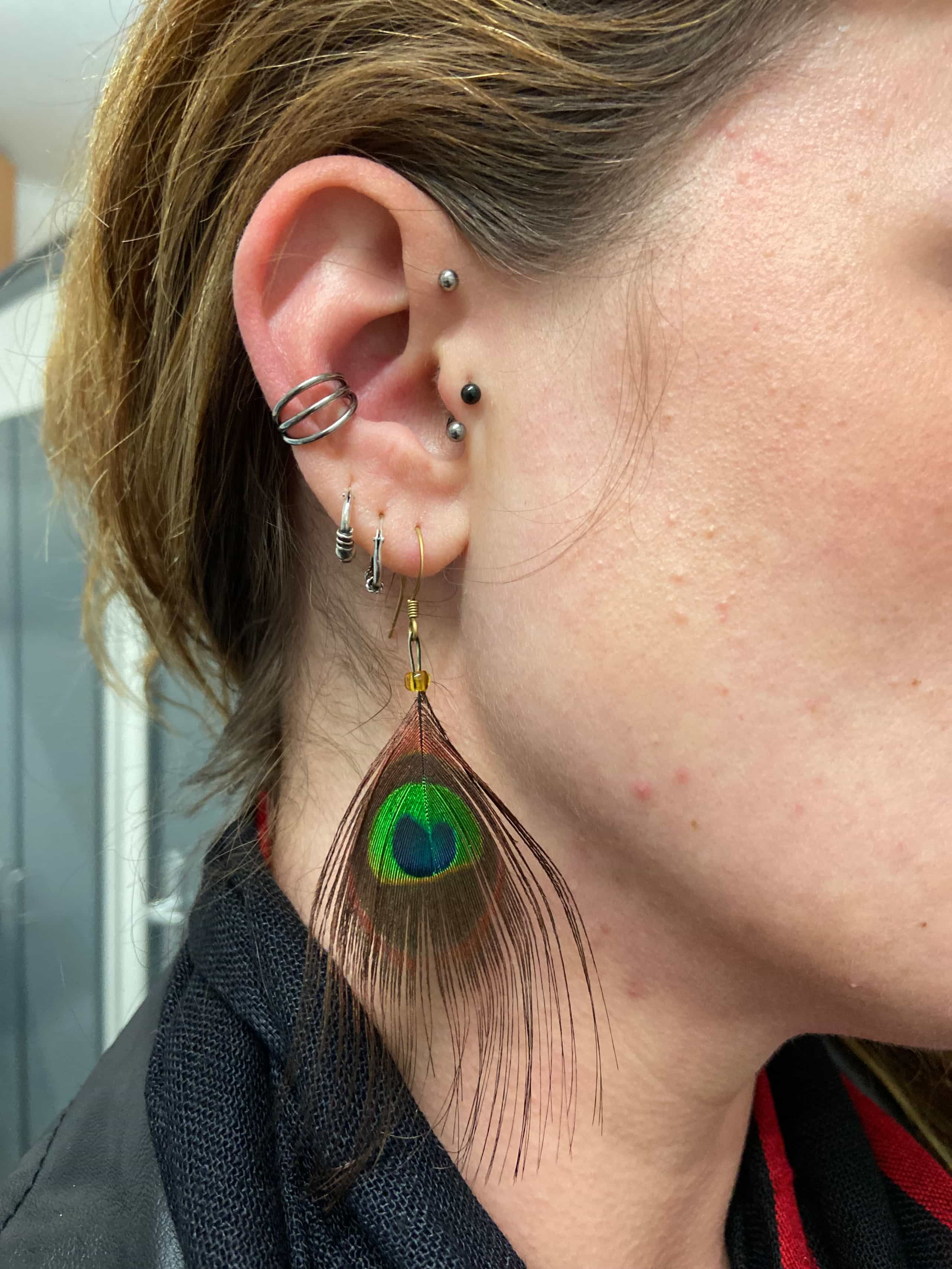 Une personne portant une boucle d'oreille en plume de paon et plusieurs piercings argentés sur l'oreille gauche, réalisés par Nadia au salon de piercing à Bastia (Bastia Tattoo).